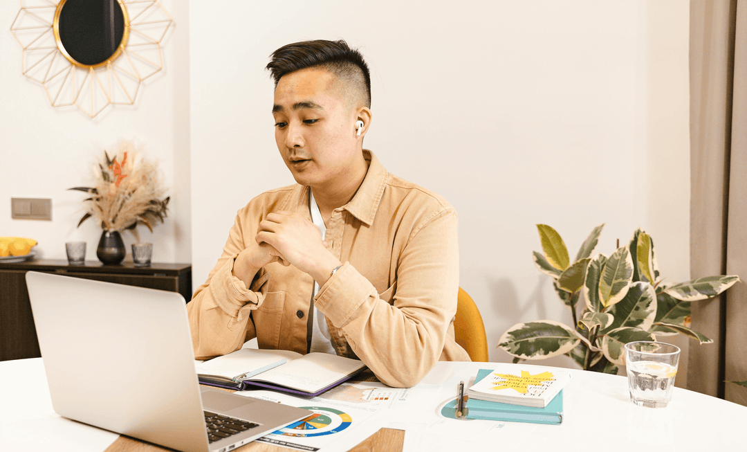 A man wearing earbuds engages in a virtual meeting at a desk with a laptop, notebooks, and a plant in the background.